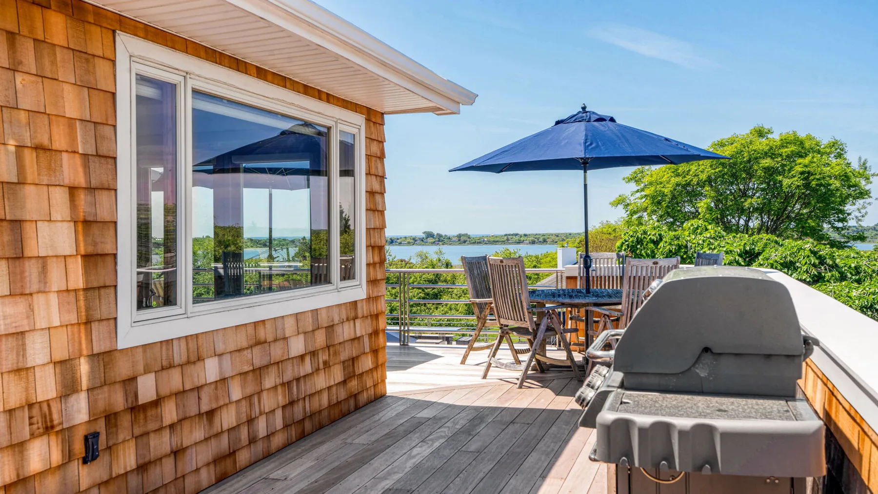An outdoor deck with dining seating, a blue umbrella, and a grill overlooks the water.