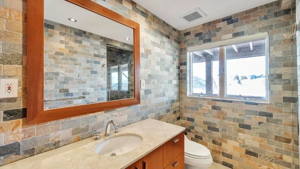 A bathroom with a stone-patterned wall, vanity, mirror, and window.