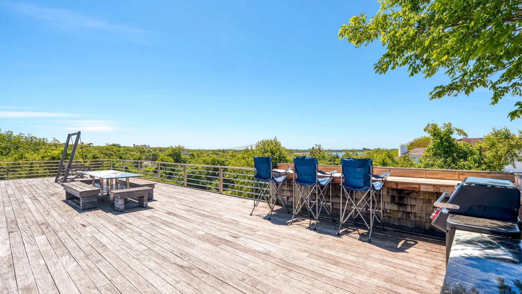 A sunny outdoor deck has a long dining table, blue chairs, and a grill.