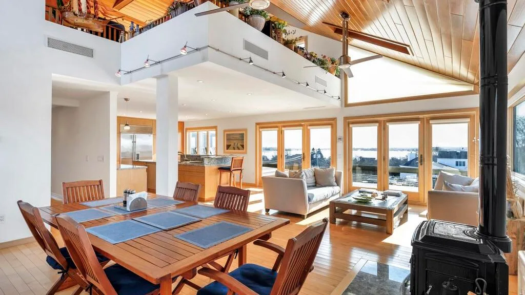 A bright open-plan living and dining area has large windows and wooden furnishings.
