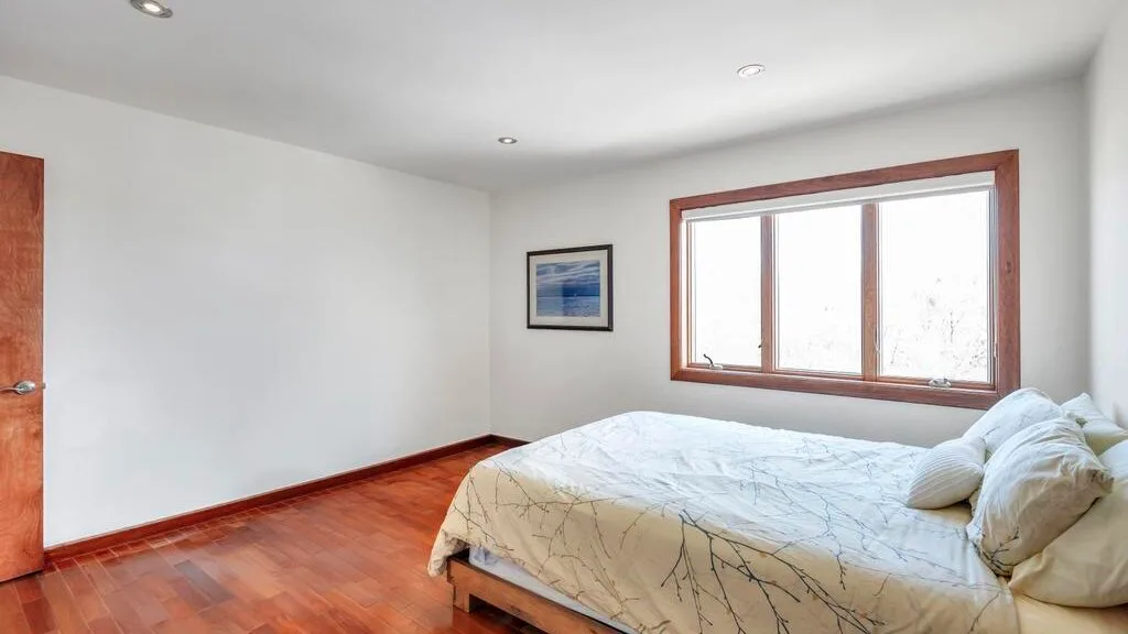 A bedroom with a bed beside a large window and wood flooring.