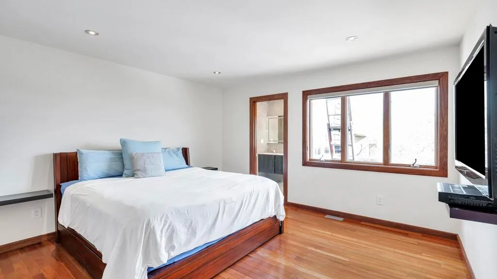 A bright bedroom with a wooden bed, white bedding, and a large window.