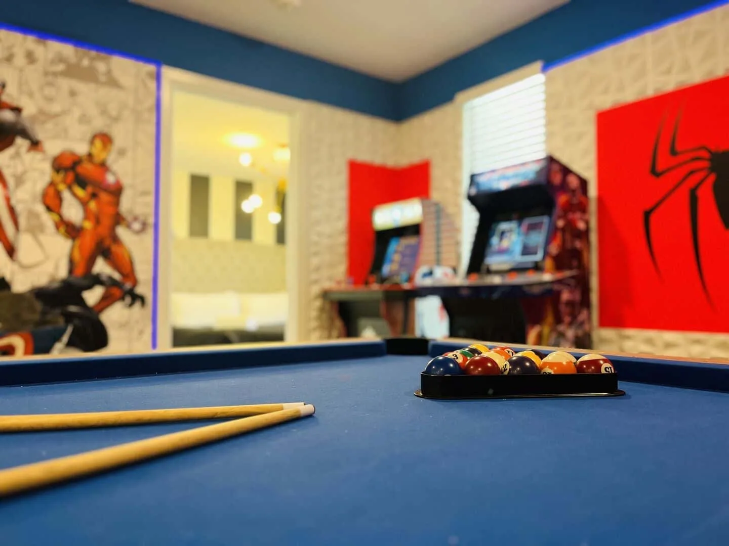 Una mesa de billar con paño azul está en una sala decorada con arte de cómics y juegos arcade.