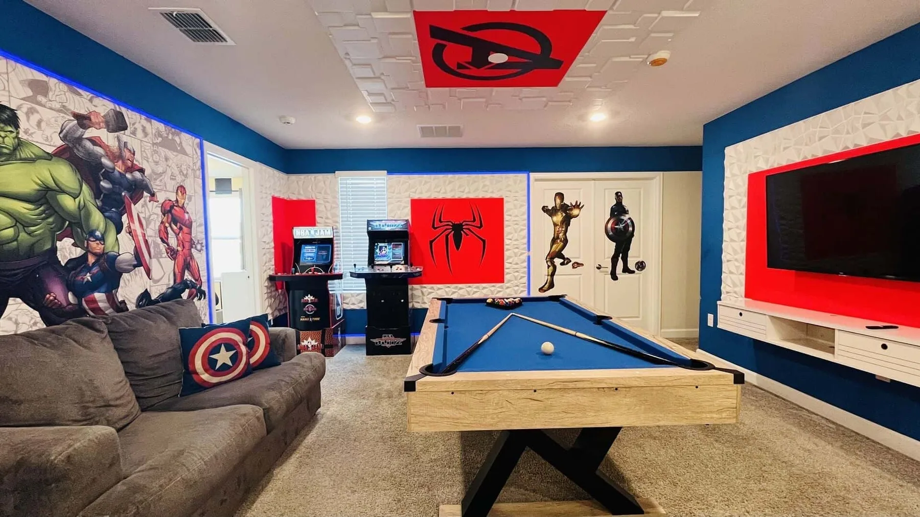 Una sala de juegos con una mesa de billar azul, asientos y arte de superhéroes en las paredes.