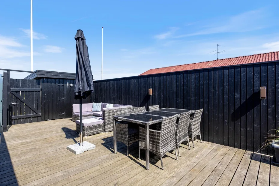 Una terraza de madera tiene una mesa de comedor exterior, asientos y una sombrilla cerrada.