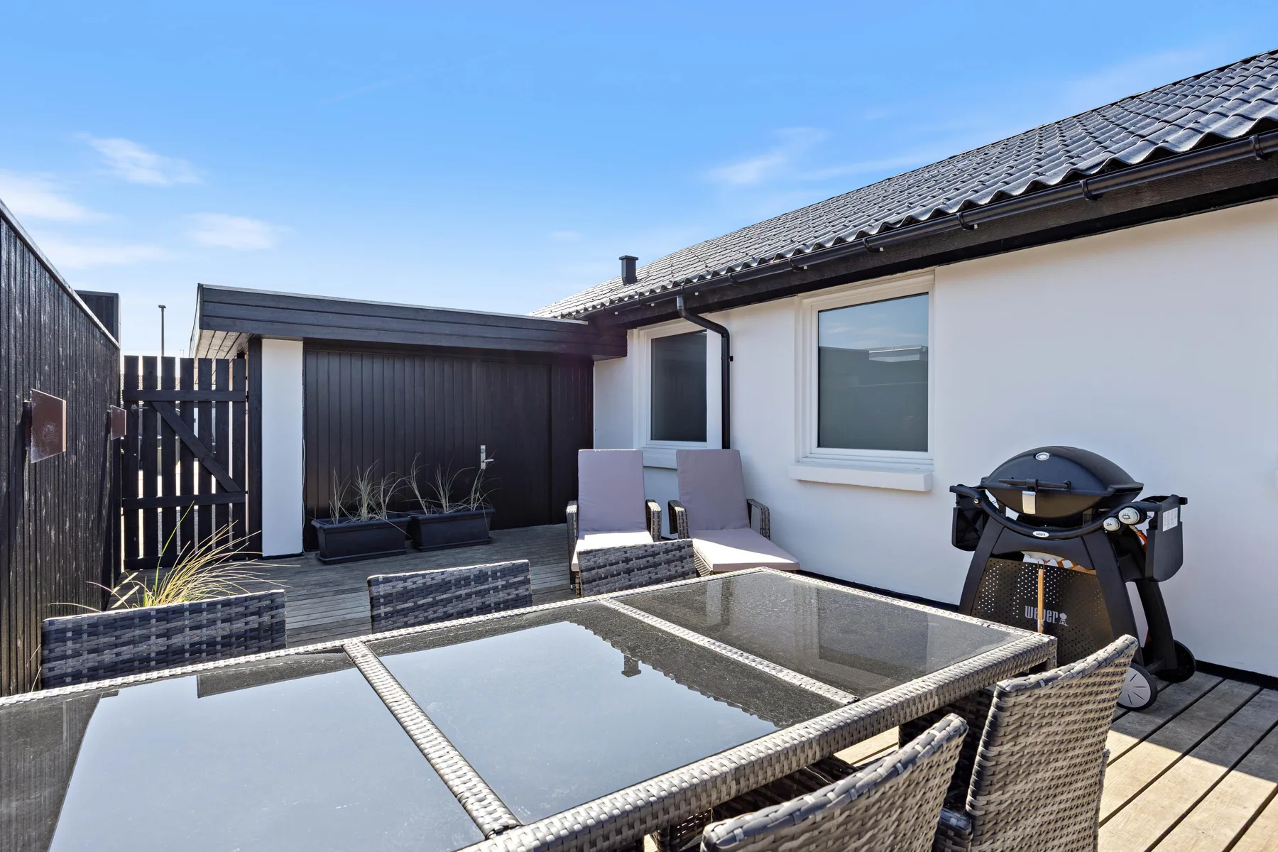 Un patio exterior tiene una mesa con tapa de vidrio, asientos y una barbacoa junto a la casa.