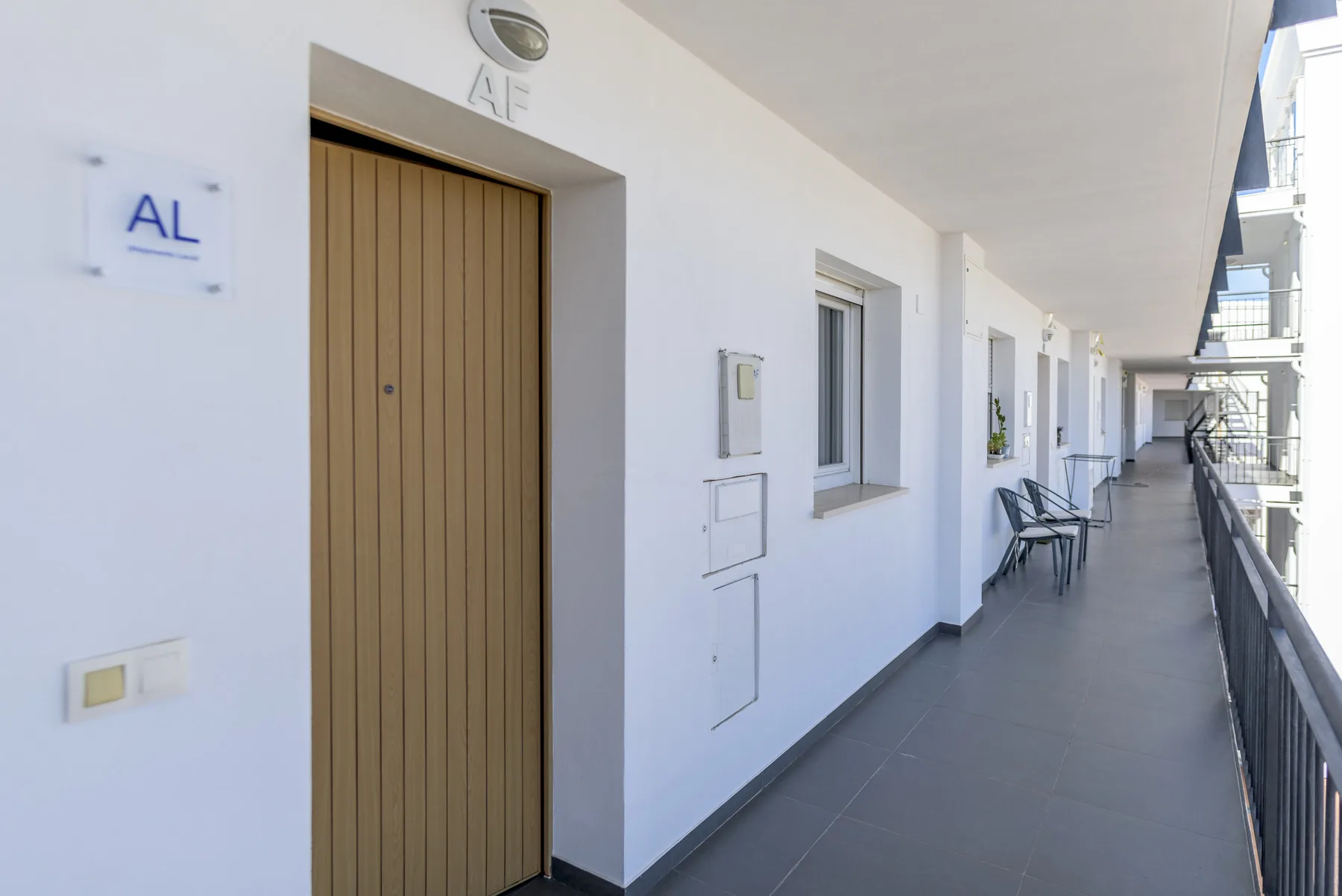 Un pasillo blanco de apartamentos con una puerta de madera y sillas junto a la barandilla.