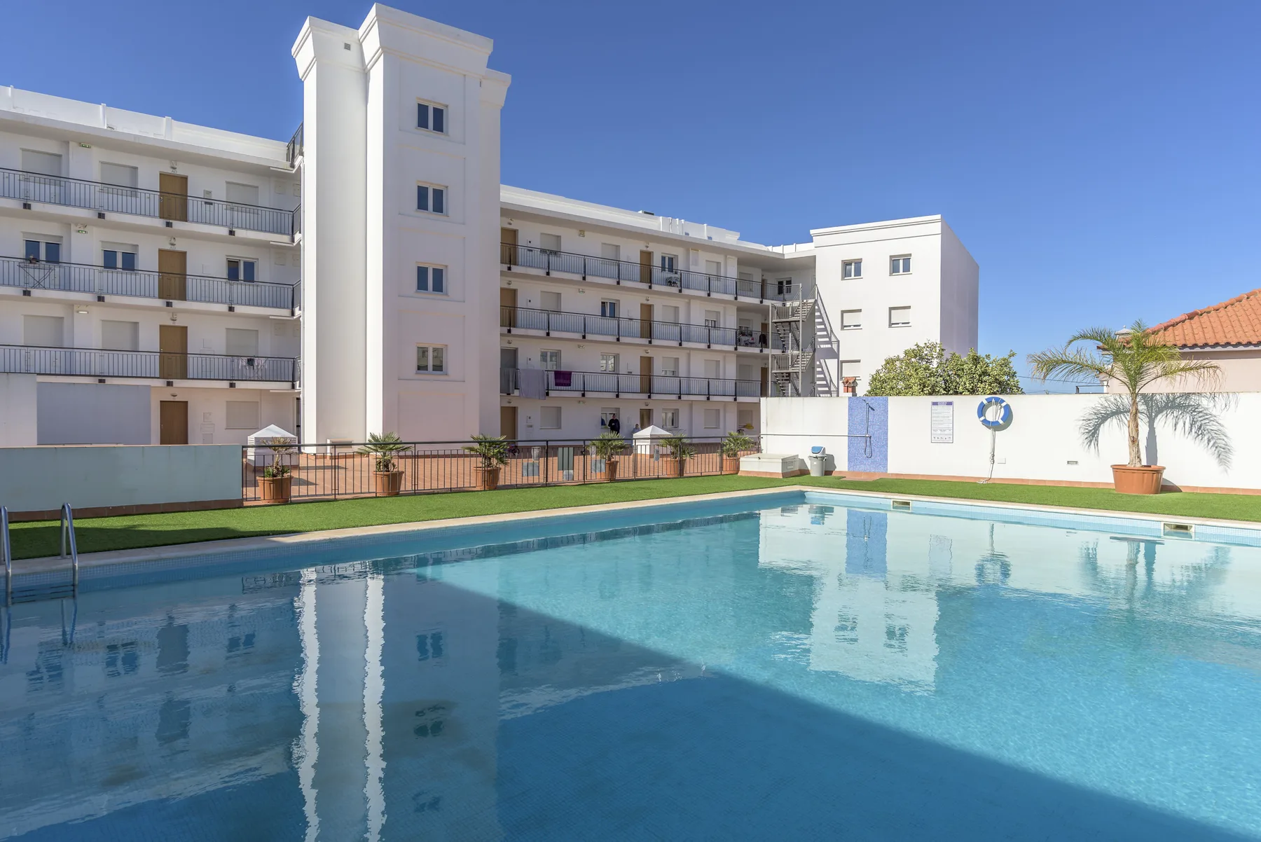 An outdoor swimming pool reflects a multi-story apartment building under a clear blue sky.