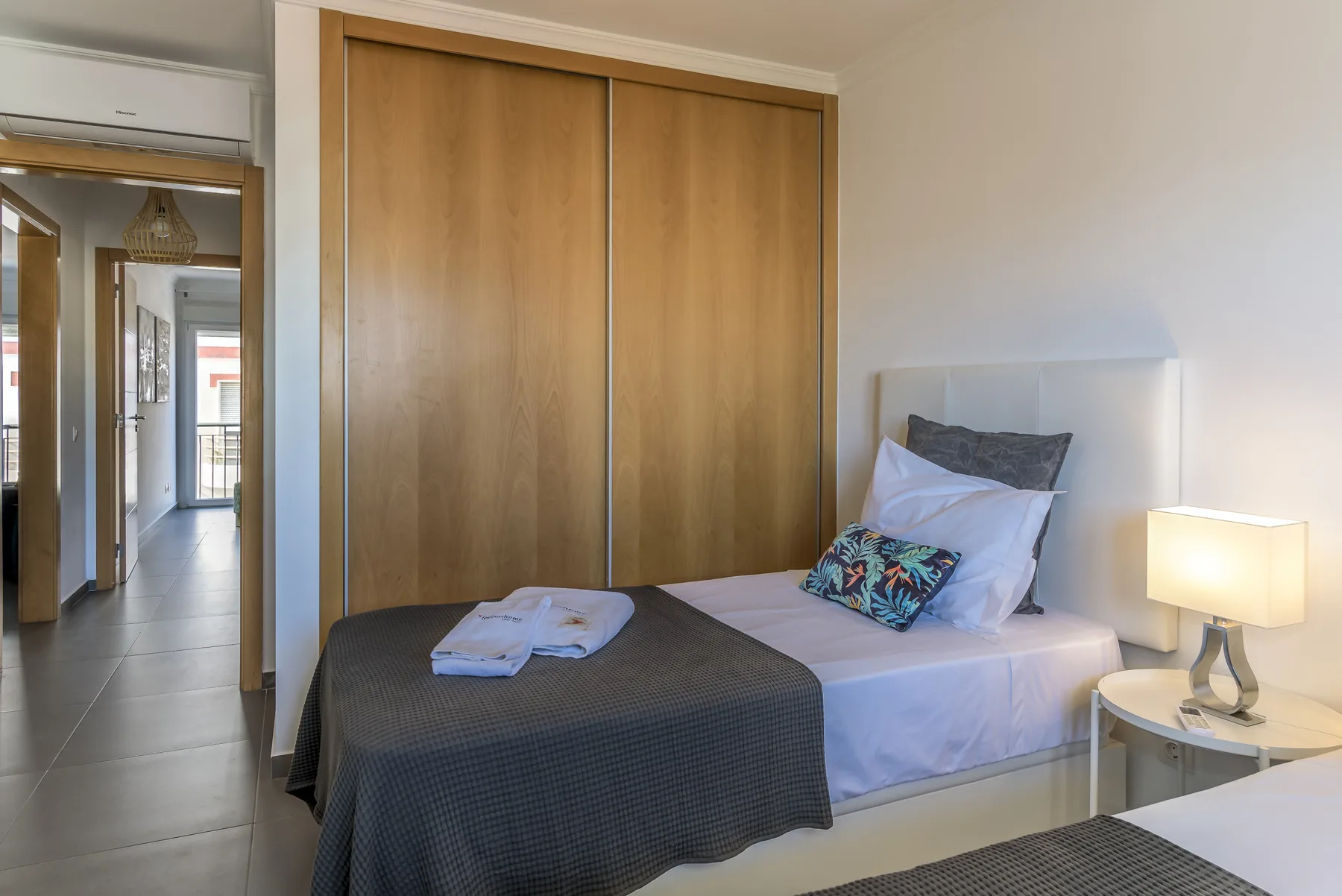 A bedroom with a bed beside a wooden wardrobe and a lit bedside lamp.