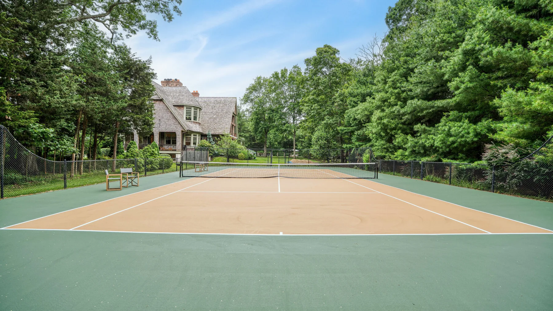 Una pista de tenis al aire libre está rodeada de árboles, con una casa al fondo.
