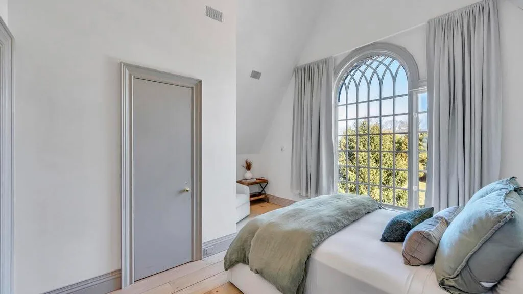 Un dormitorio con una cama junto a una ventana alta en arco y cortinas.