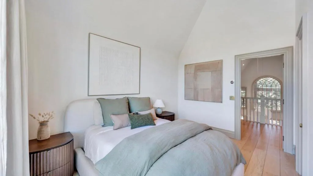 Un dormitorio luminoso con una cama bien tendida y una puerta hacia un pasillo.