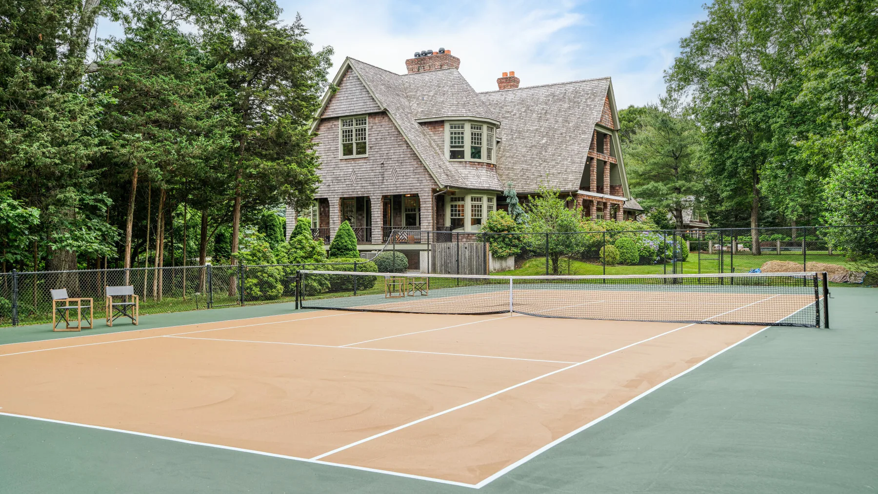 Una pista de tenis al aire libre está frente a una casa grande rodeada de árboles.