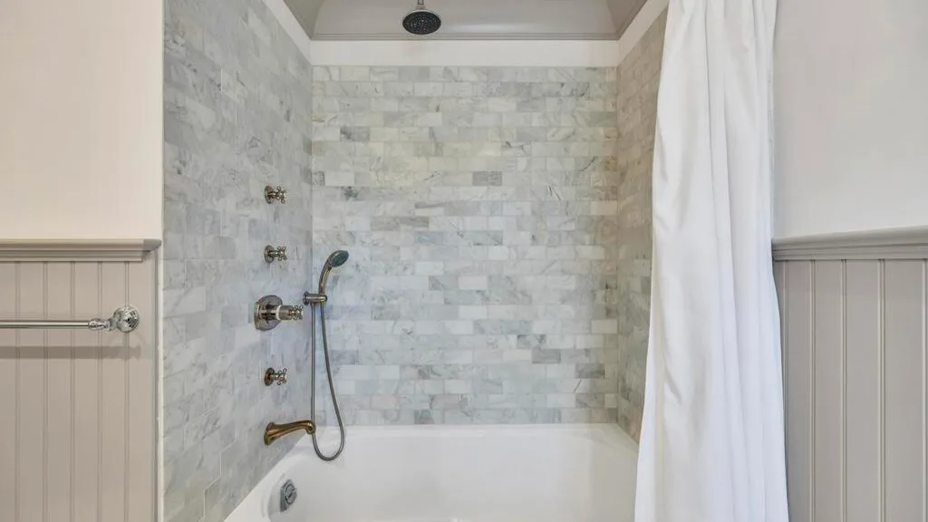 Zona de ducha de baño con azulejos, bañera y cortina blanca.