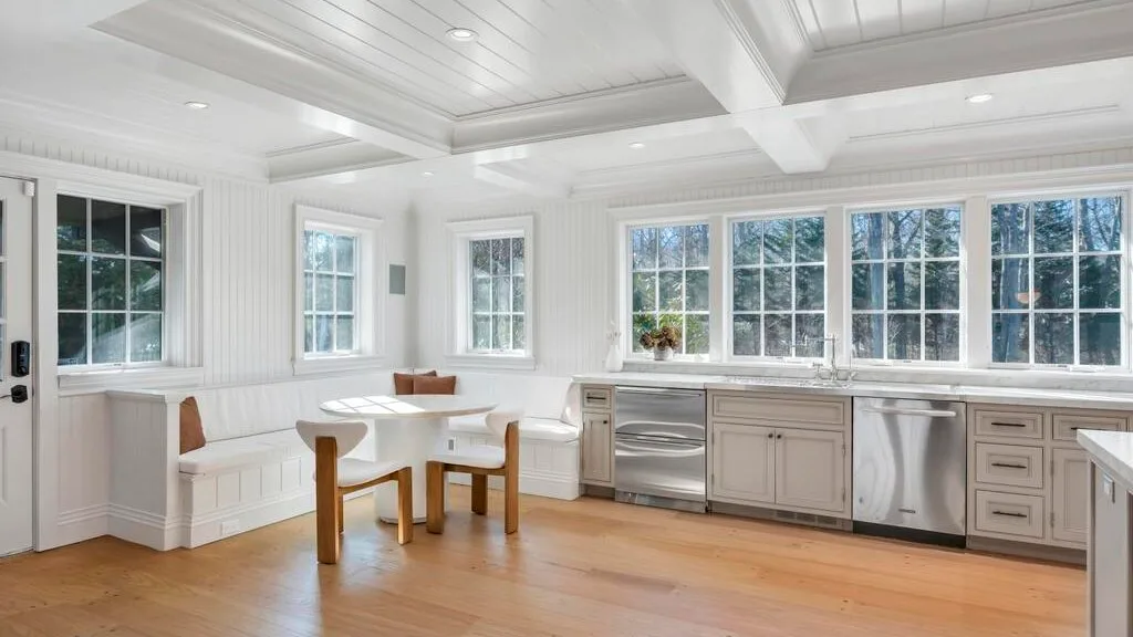 Una cocina luminosa con gabinetes blancos, ventanas grandes y una mesa pequeña.