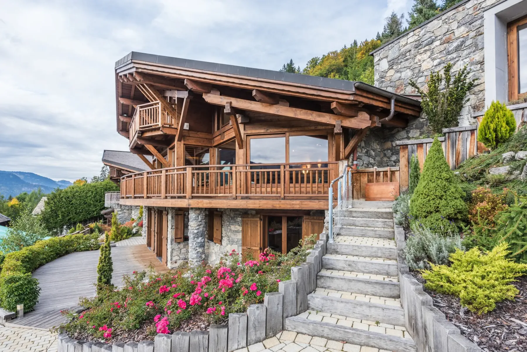 Vista exterior de un chalet con balcón de madera, muros de piedra y escaleras en el jardín.
