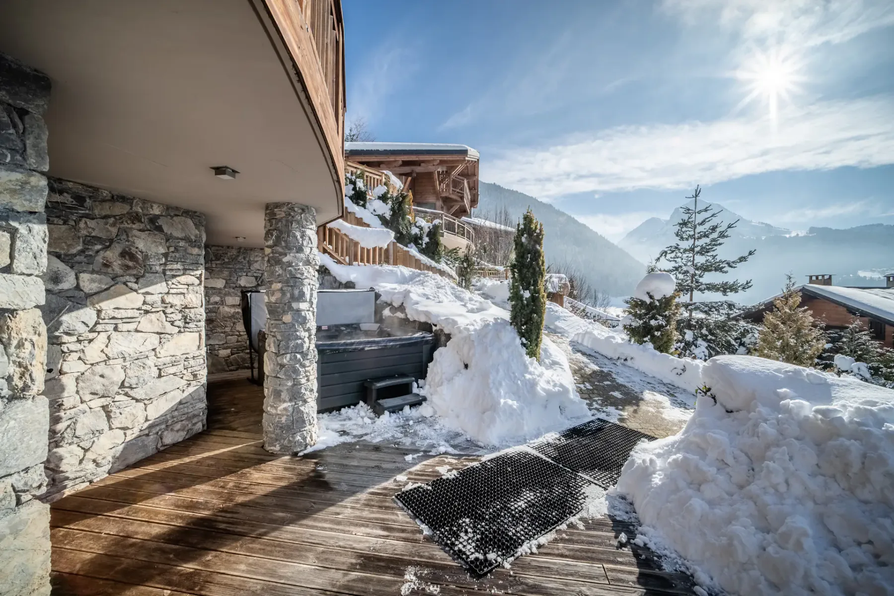 Un jacuzzi está en una terraza de madera nevada junto a un edificio de piedra, con vistas a las montañas.