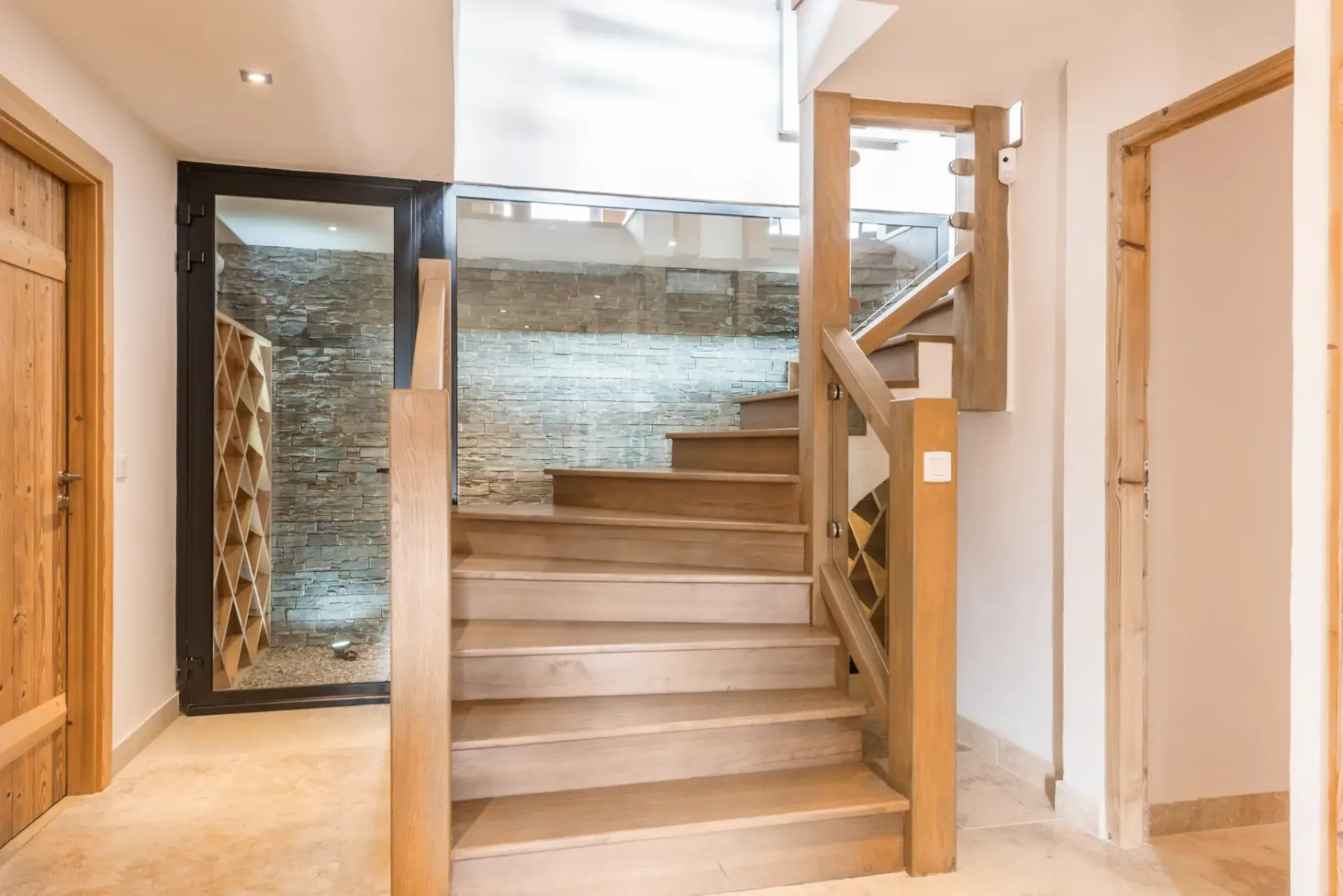 Una escalera de madera sube junto a una puerta de cristal en un interior luminoso.