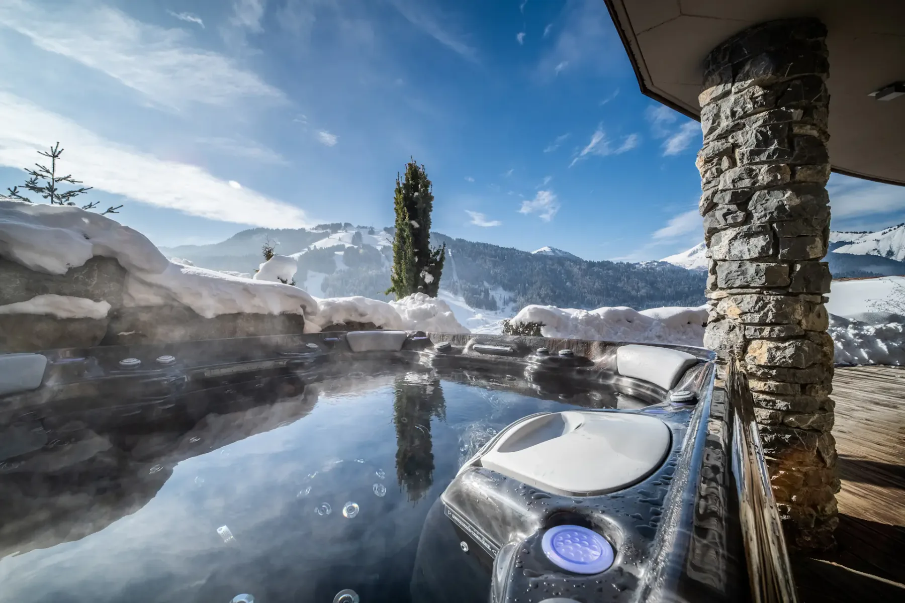 Una bañera de hidromasaje exterior está junto a muros de piedra cubiertos de nieve, con montañas al fondo.