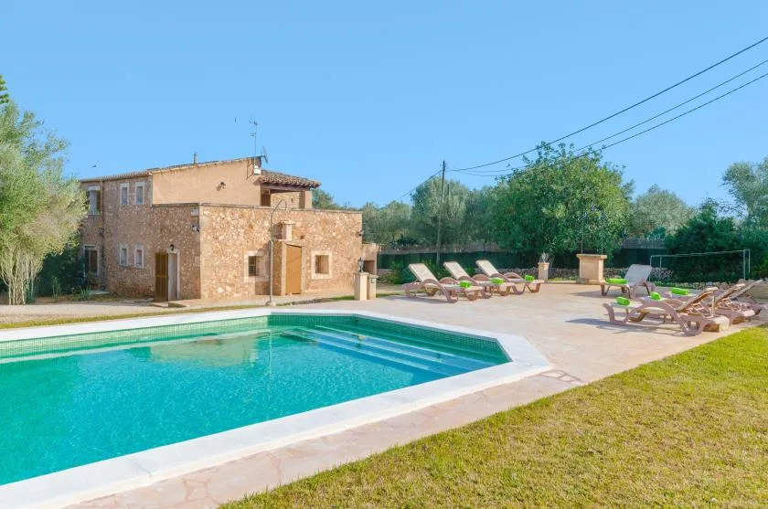 Una piscina al aire libre junto a una casa de piedra, con asientos en la terraza pavimentada.
