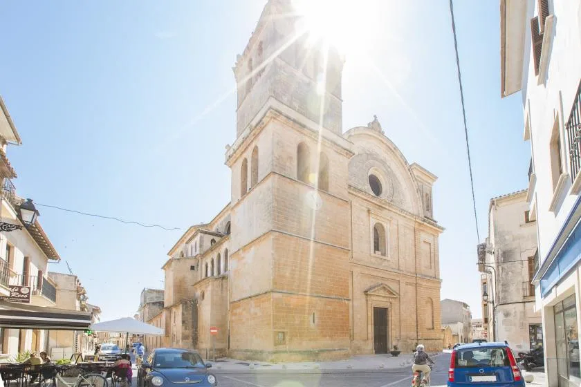 Una gran iglesia de piedra está junto a una calle soleada con coches estacionados.