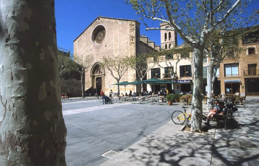 A sunny public square with a stone church, café seating, and leafy trees.