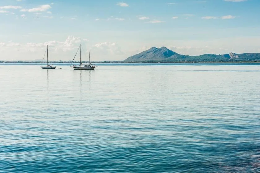 A calm bay with two sailboats and distant mountains beneath a partly cloudy sky.