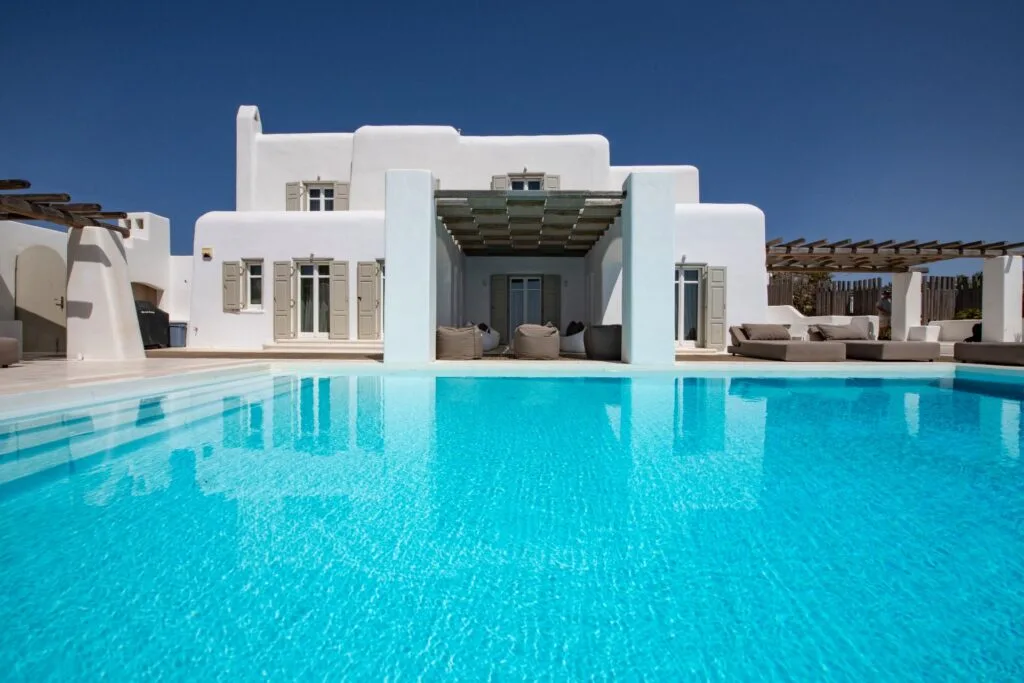 A turquoise swimming pool sits in front of a white villa with shaded seating.