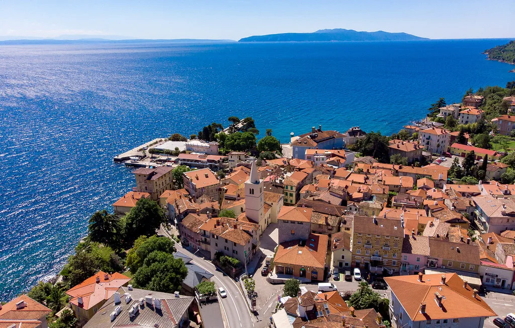 Aerial view of a coastal town with red-tiled roofs beside blue water.