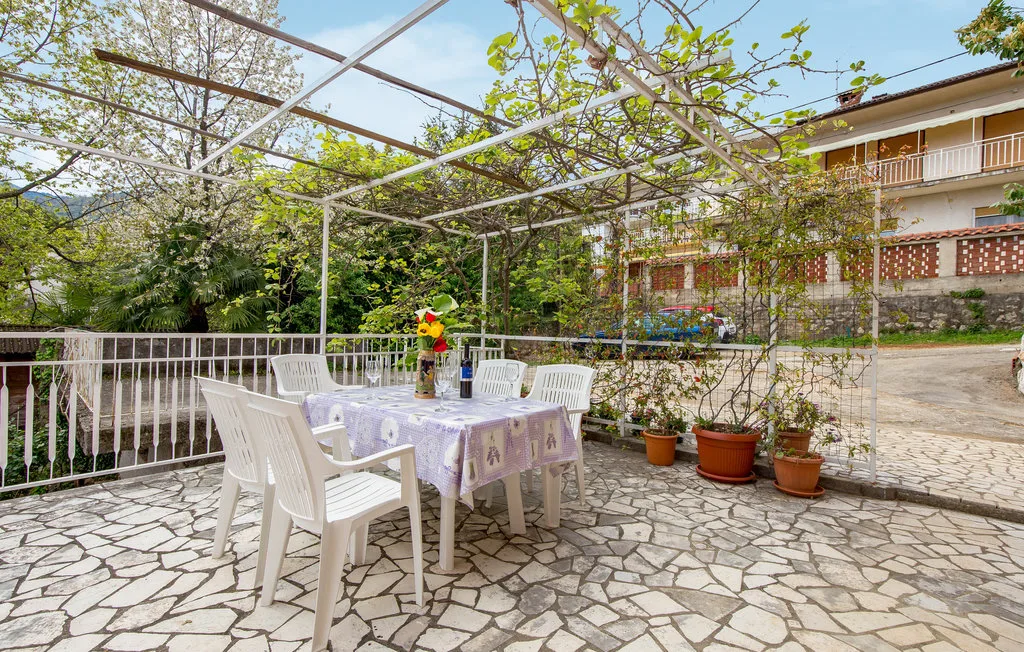 A stone-paved outdoor dining area with a table and white seating beneath a vine-covered pergola.