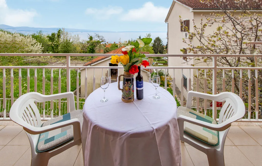 A balcony dining table with flowers and seating overlooks neighboring buildings and greenery.