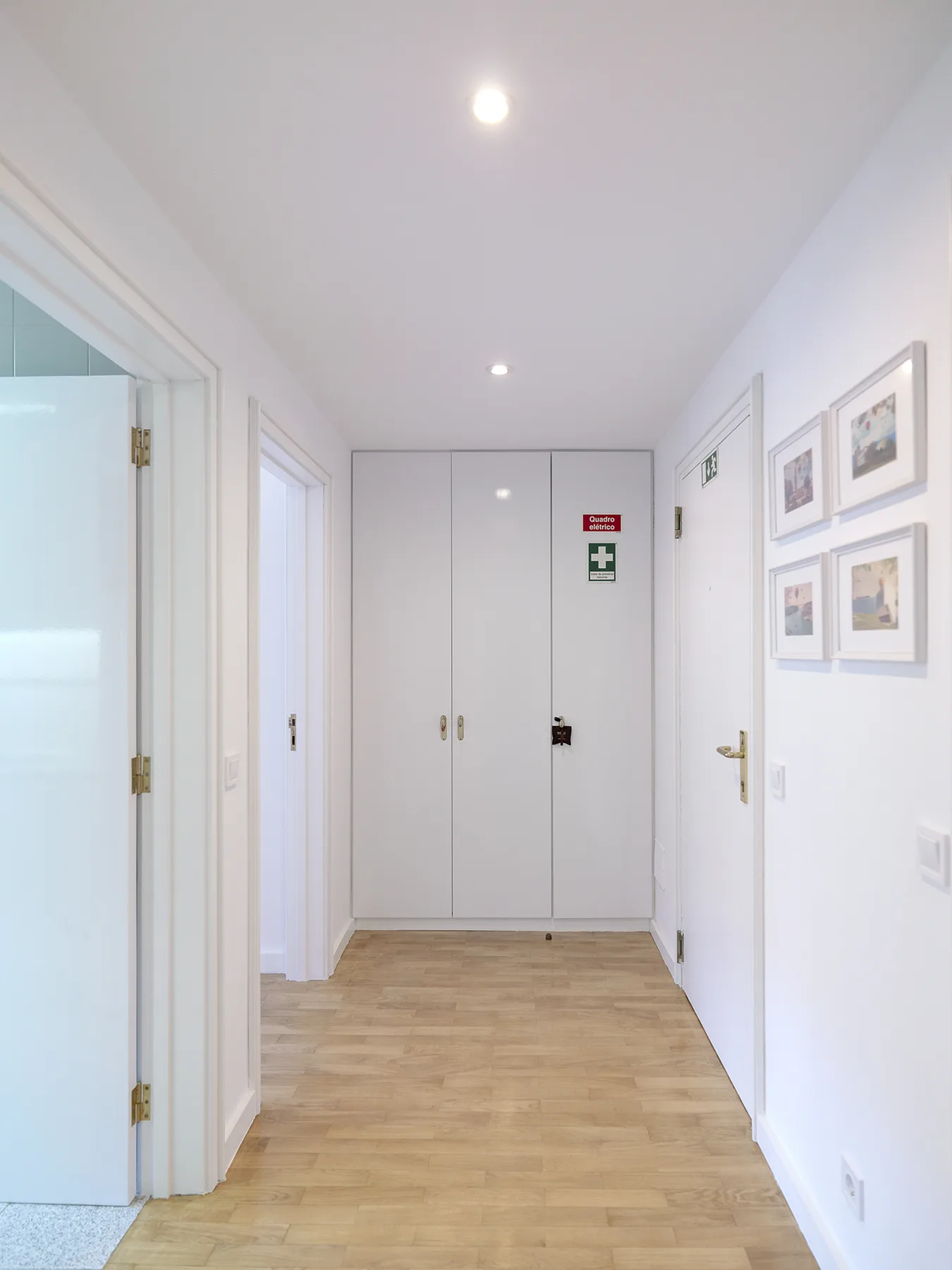 Un pasillo luminoso con suelo de madera clara, cuadros enmarcados y puertas blancas.