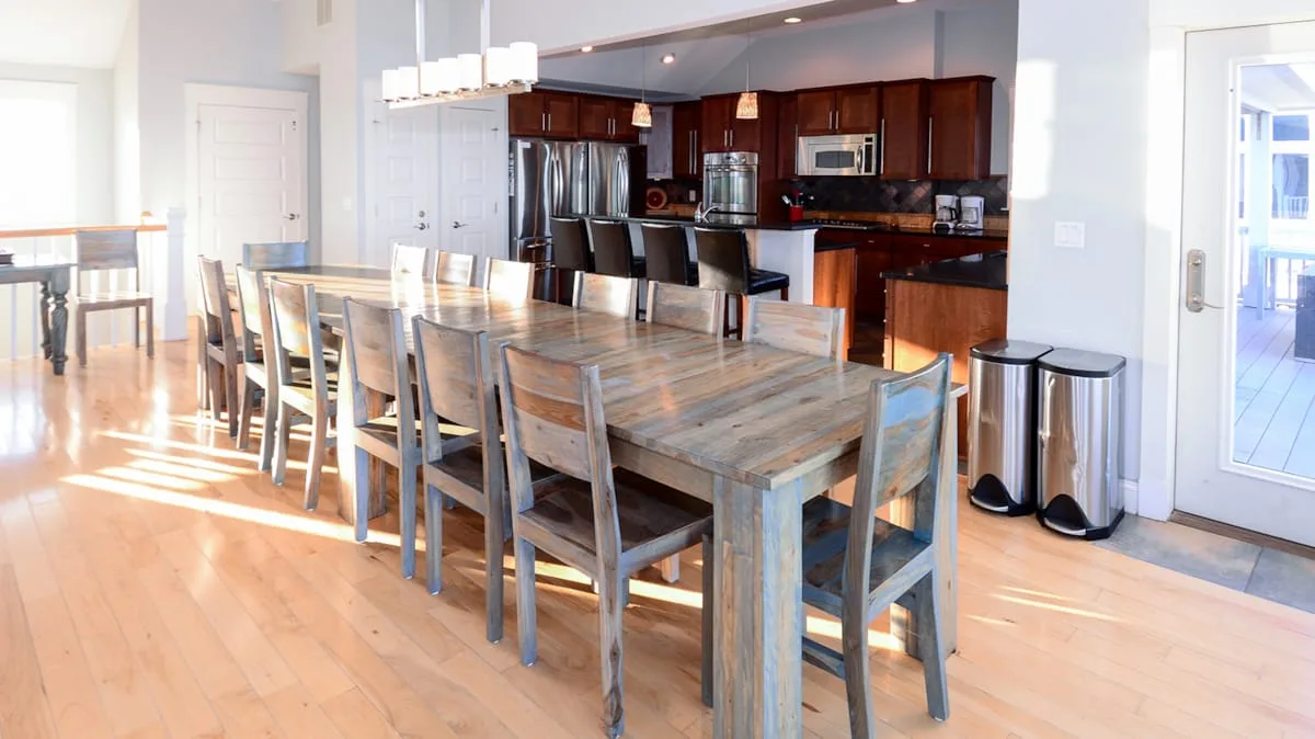 Comedor con una mesa larga de madera y gabinetes oscuros de cocina al fondo.