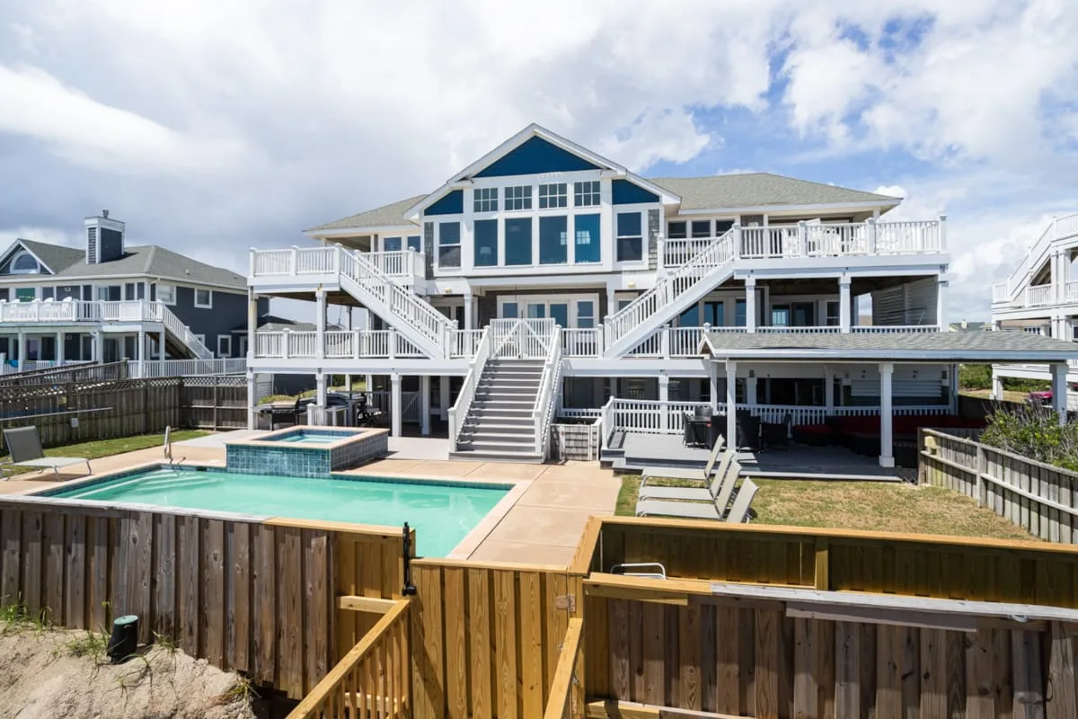 A large coastal-style house overlooks a fenced outdoor pool and wooden deck.