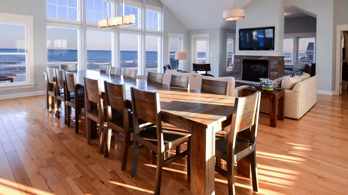Comedor iluminado junto a grandes ventanas con vistas al agua y una larga mesa de madera.
