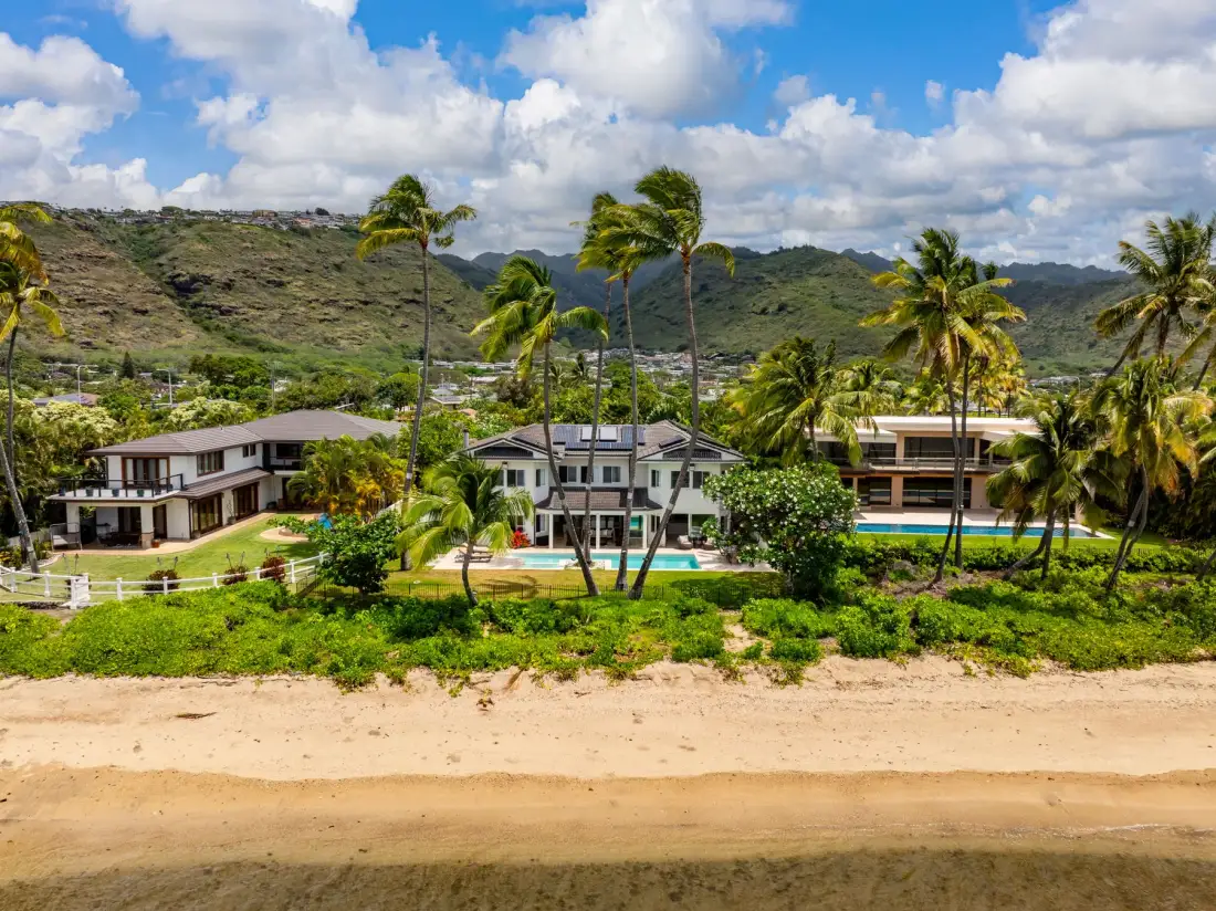A beachfront resort with palm trees, swimming pools, and green hills in the background.