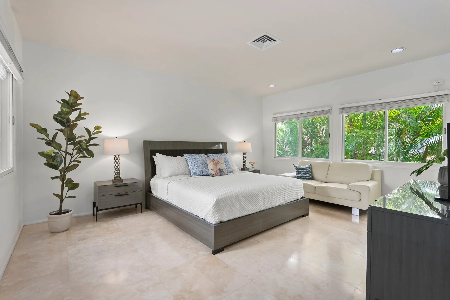 A bright bedroom with a large bed, a seating area, and windows overlooking greenery.