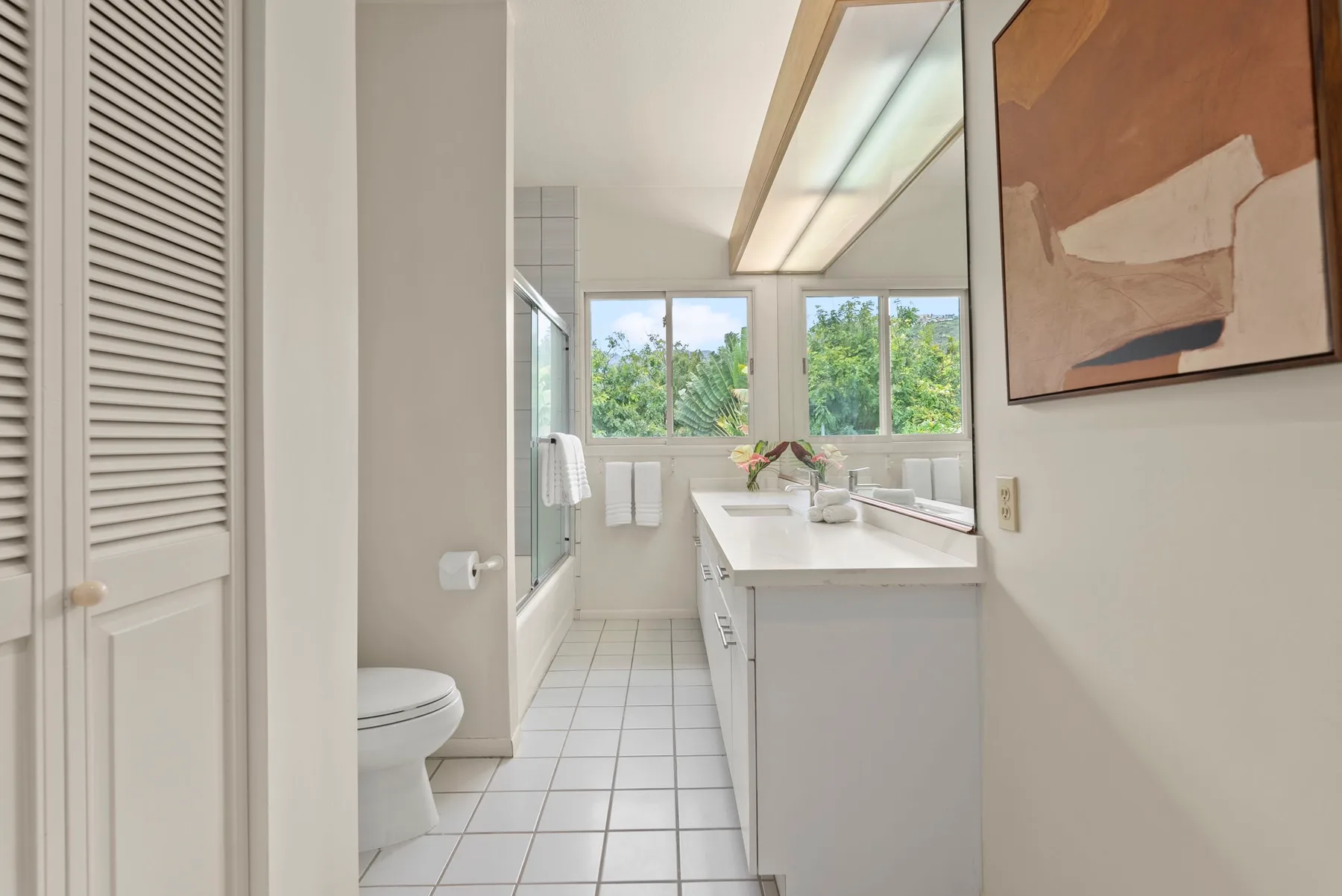 A bright bathroom with a long vanity, tiled floor, and windows overlooking greenery.