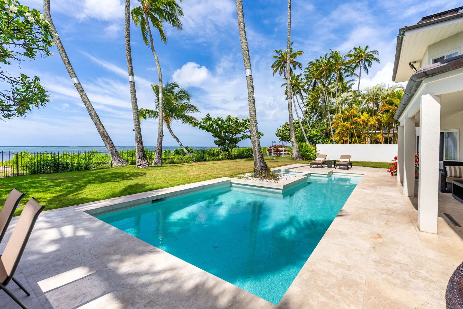 An outdoor swimming pool beside a white building, with palm trees and the ocean beyond.