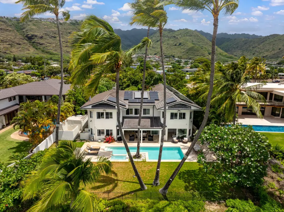 Aerial view of a white house with a swimming pool, palm trees, and green hills.