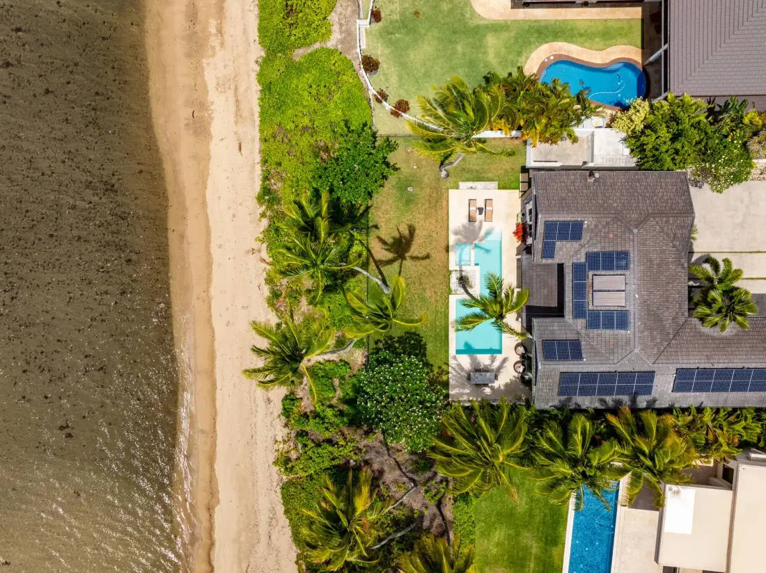 An aerial view shows a beachfront property with a house, green lawn, and swimming pools.