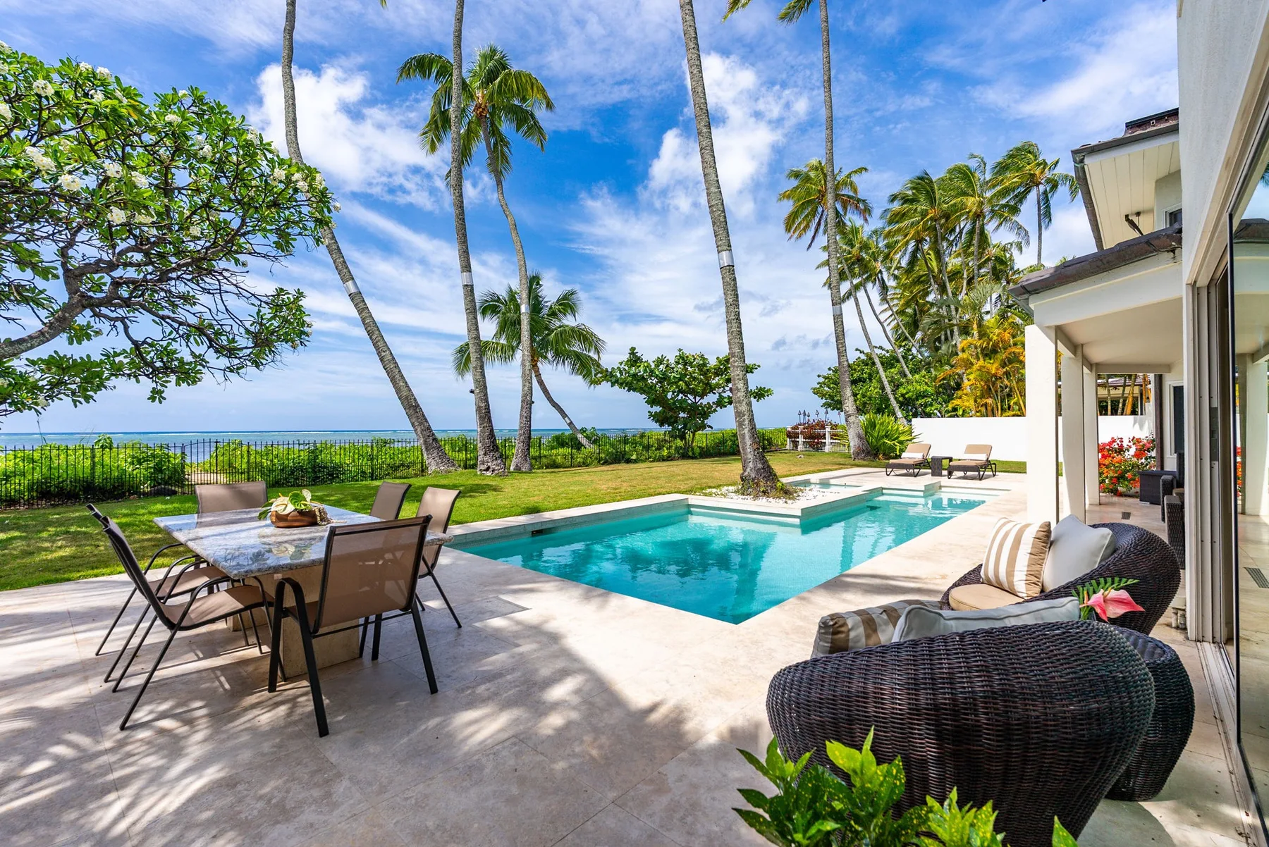 Un patio tropical con piscina, asientos al aire libre y palmeras frente al océano.