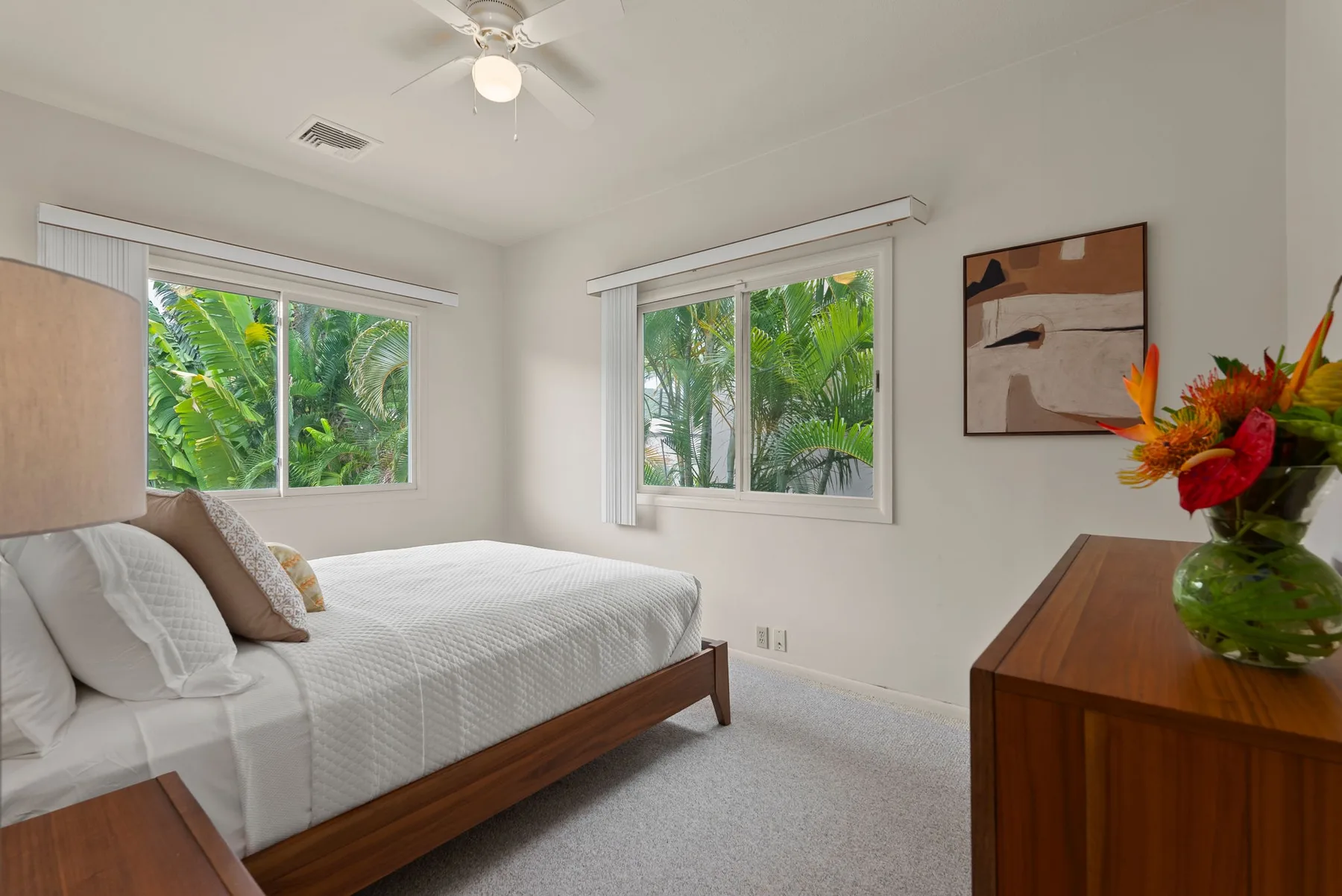 A bright bedroom with a bed, windows, and leafy greenery outside.