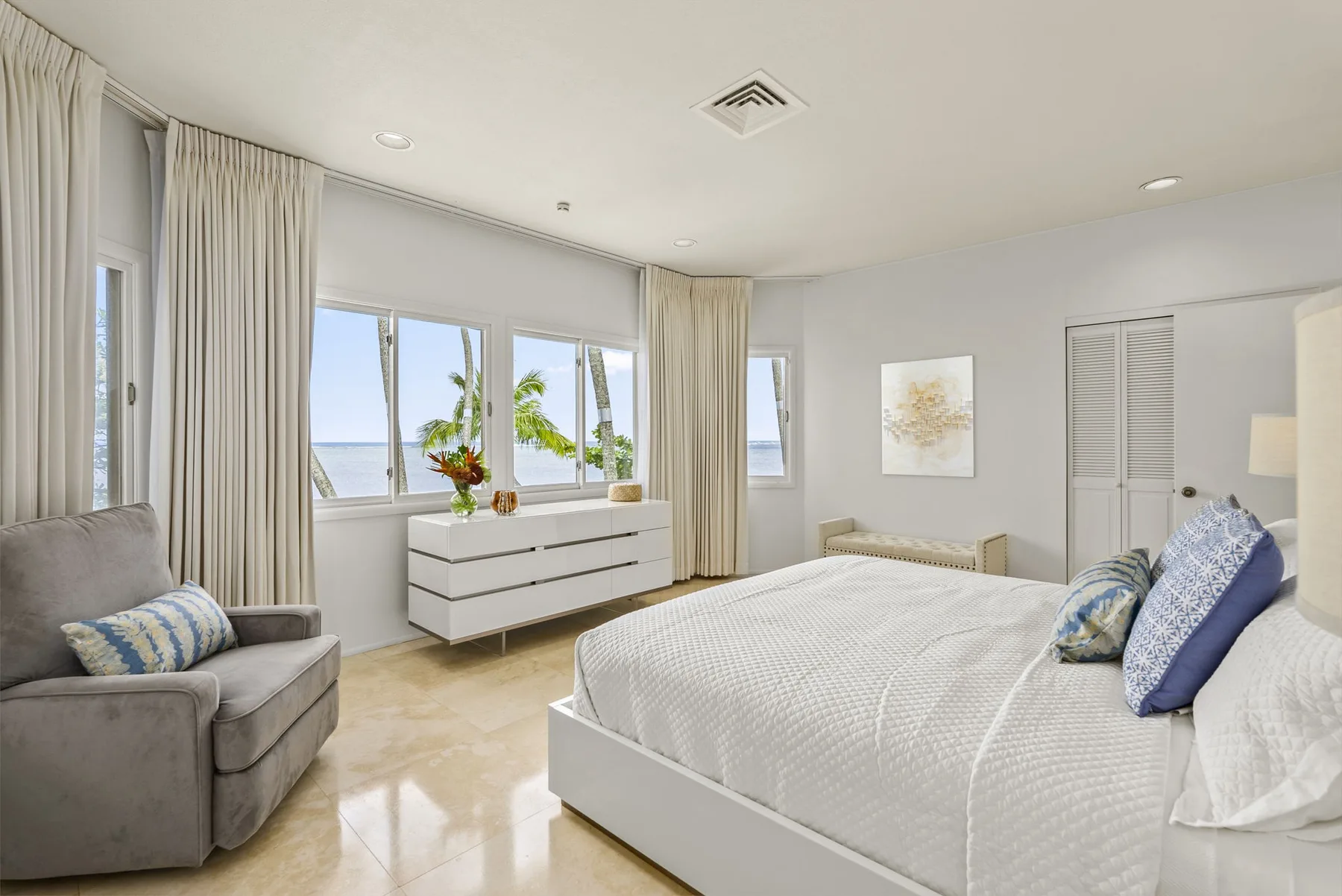 A bright bedroom with a bed, an armchair, and windows overlooking the water.