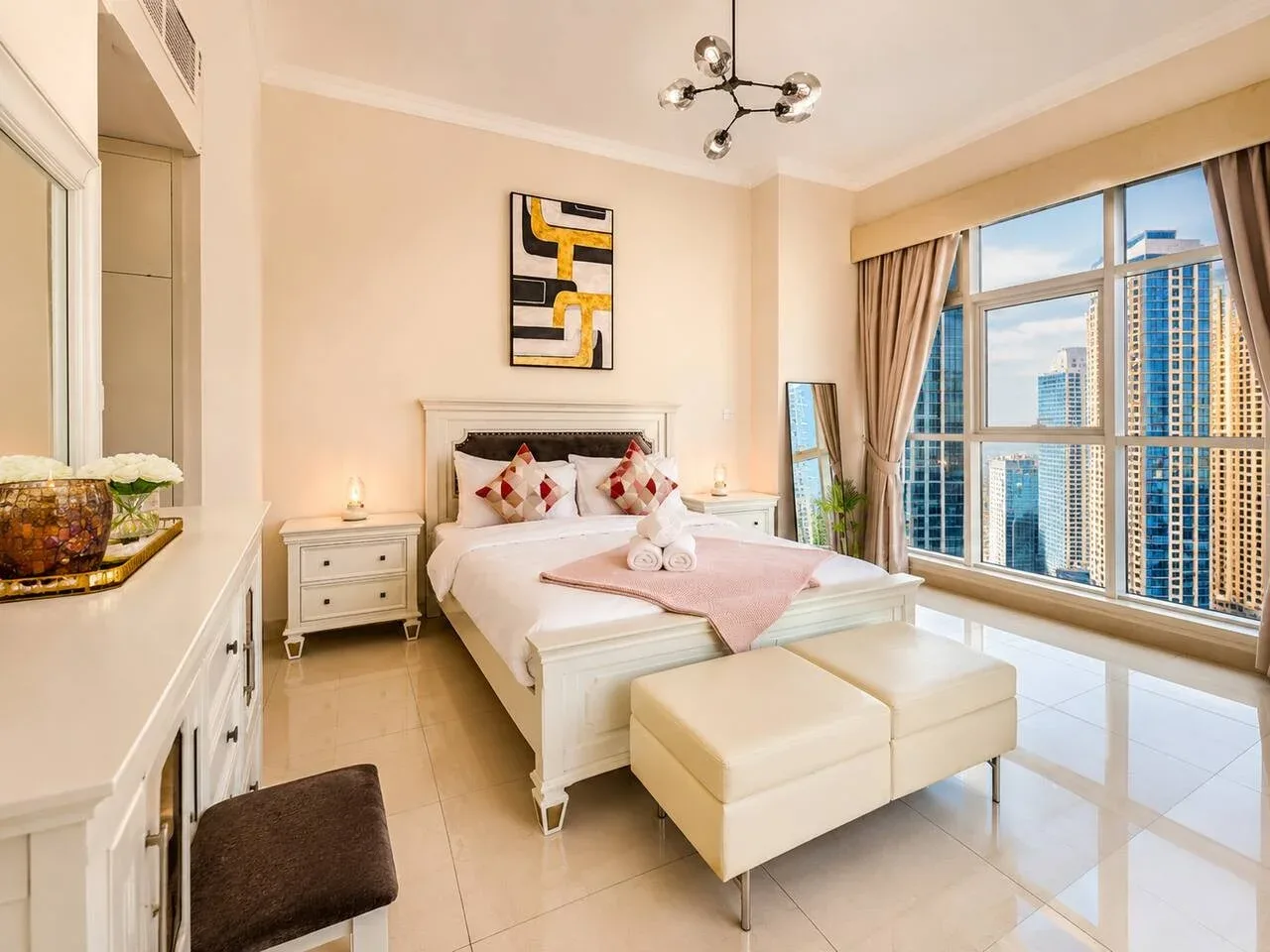 A bright bedroom with a bed, seating, and large windows overlooking nearby buildings.
