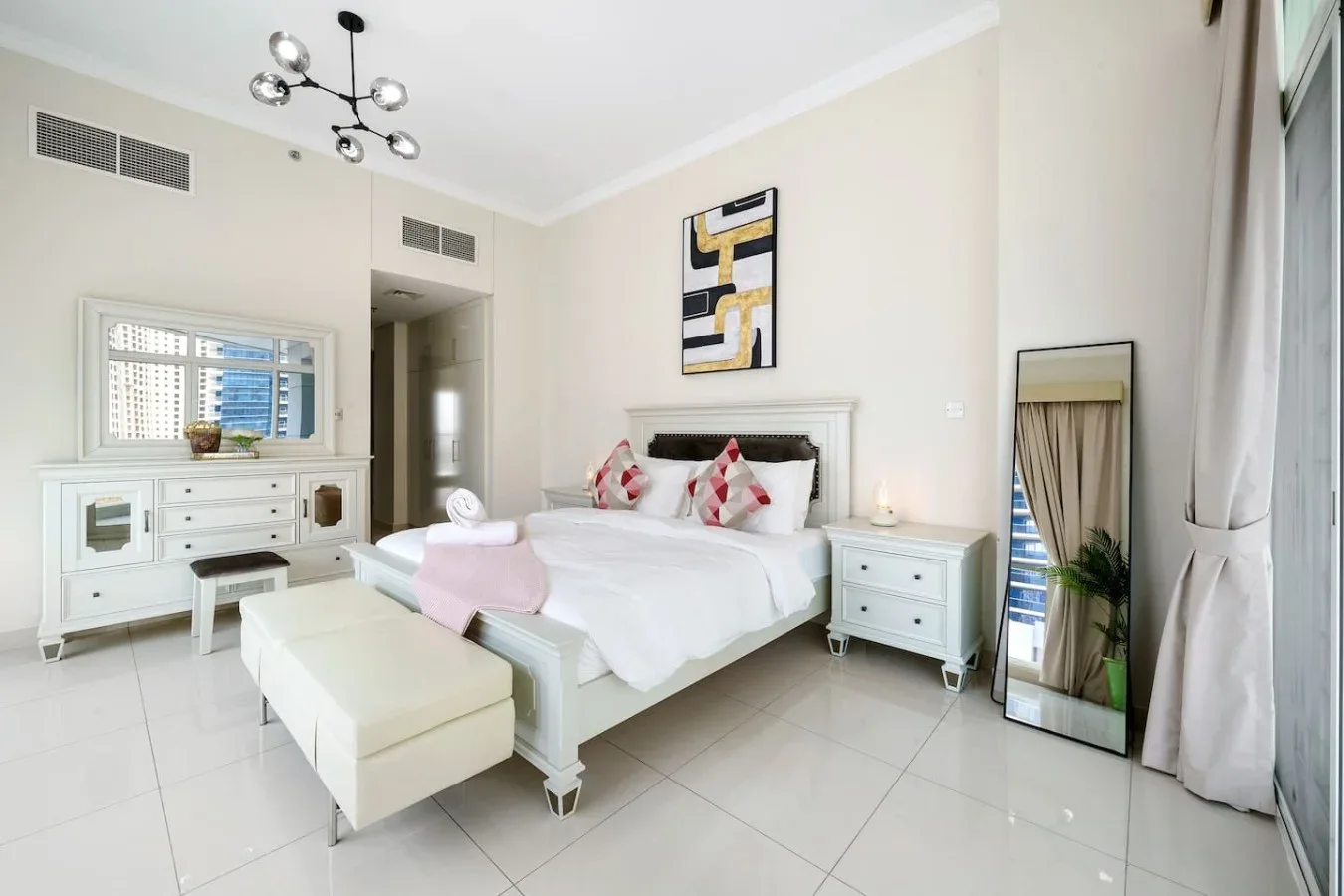 A bright bedroom with a bed, matching bedside tables, and a full-length mirror.