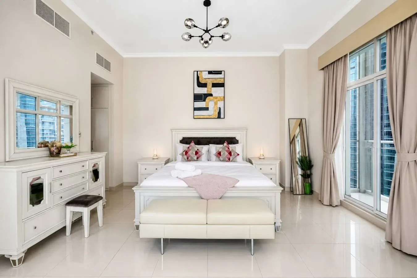 Un dormitorio luminoso con una cama, muebles claros y ventanas altas con cortinas.