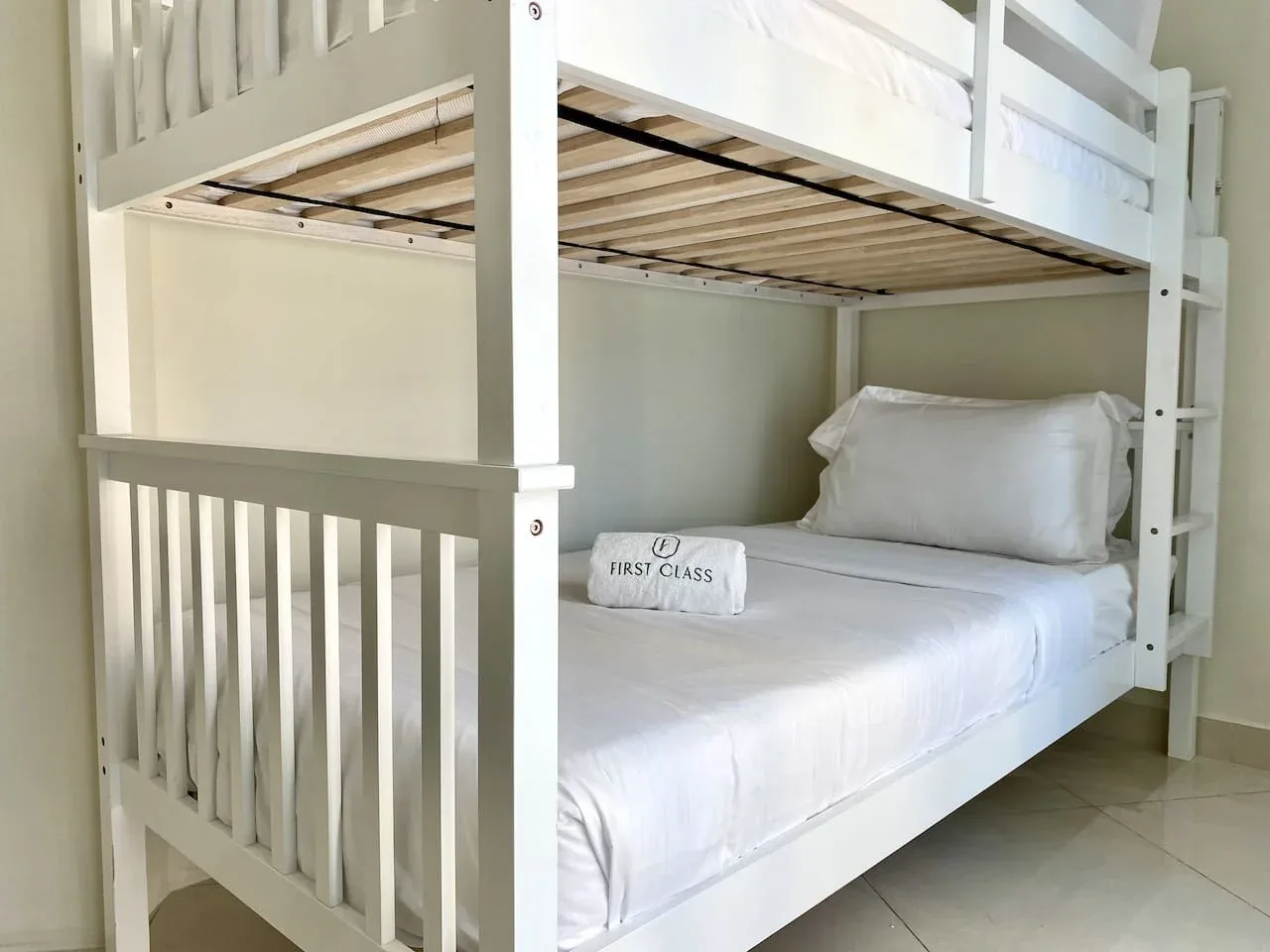 A white bunk bed with neatly made bedding sits against a light-colored wall.