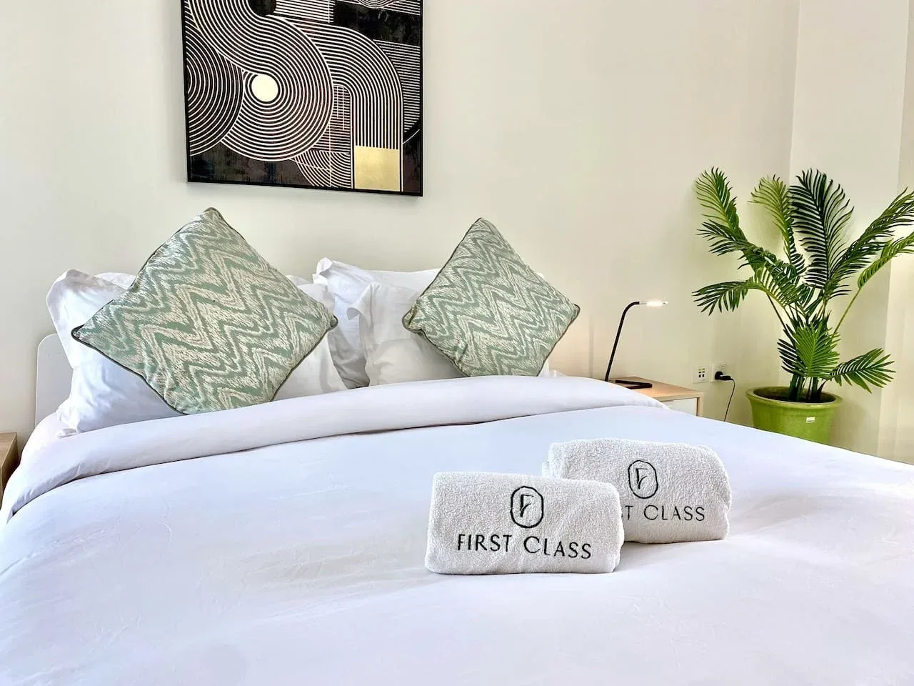 A neatly made bed with patterned pillows and folded towels beneath abstract wall art.