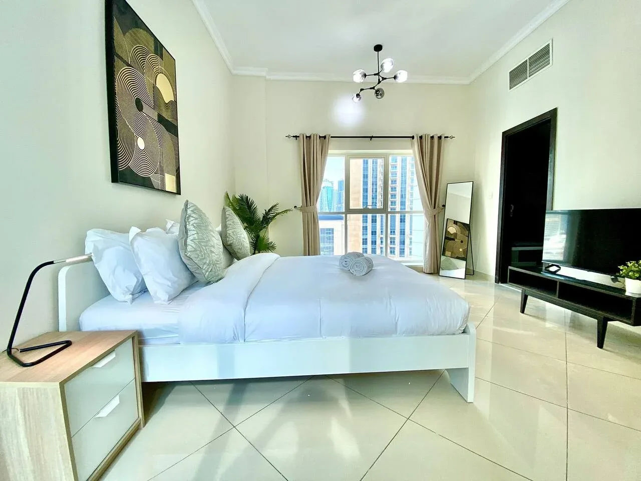 A bright bedroom with a bed facing a television and a large curtained window.